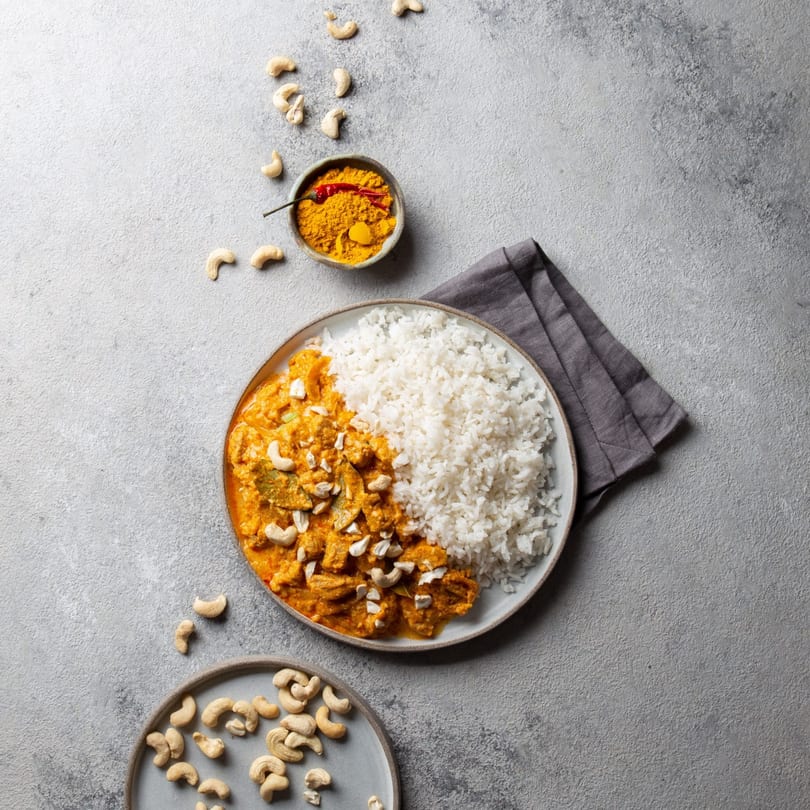 Indian chicken curry with rice, cashews, and a bowl of turmeric and chili on a gray surface.