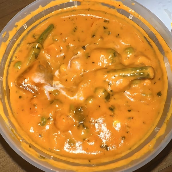 Close-up of a vibrant orange Indian curry with vegetables and a swirl of cream in a plastic container.